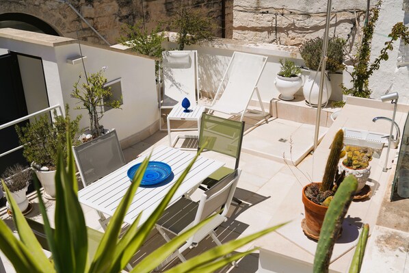 Un patio soleado con sillones blancos, una mesa con bandeja azul y diversas plantas en macetas.