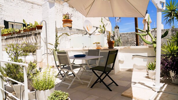 Un patio exterior con mesa y sillas, rodeado de plantas en macetas y una pared blanca.