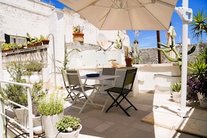 Une terrasse extérieure avec une table et des chaises, entourée de plantes en pot et d’un mur blanc.