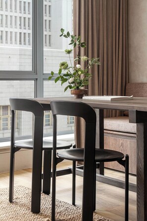 Ein moderner Essbereich mit Holztisch und schwarzen Stühlen, einer Topfpflanze und Blick auf eine Stadtlandschaft durch große Fenster.