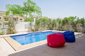A swimming pool with two bean bags, surrounded by greenery and a building in the background. - Vesta - Luxury Villa - 4BR - CFC (New Cairo)