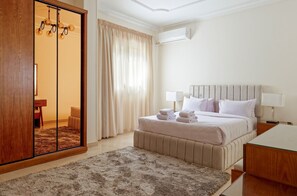 A hotel room with a bed, bedside lamps, a wooden wardrobe, a mirror, and a view of the bathroom. - Vesta - Luxury APT - 3BR - New Cairo IV (New Cairo)