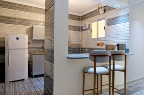 A compact kitchen with a refrigerator, a microwave, and a small countertop. There are two bar stools and a window above the countertop. - Vesta - Luxury APT - 3BR - New Cairo IV (New Cairo)