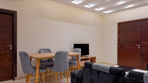 A room with a wooden dining table, grey upholstered chairs, a black flat-screen TV, and a dark grey sofa. - Vesta - Luxury APT - 2BR - New Cairo II (New Cairo)