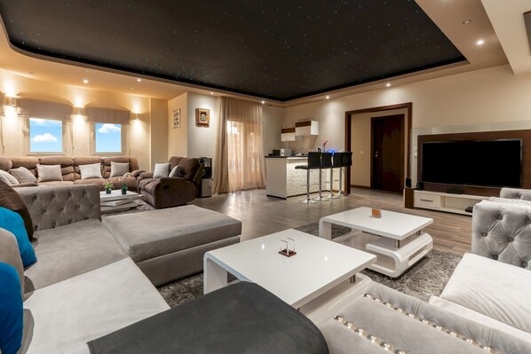 A modern living room with a sectional sofa, a flat-screen TV, and a kitchen area visible through an open door. - Vesta - Luxury Villa - 6BR - New Cairo (New Cairo)