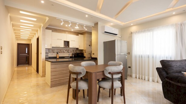 A modern kitchen with a central island, a dining area with four chairs, and a living room with a sofa. - Vesta - Luxury APT - 2BR - Kattameya one (Cairo)
