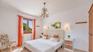 Un dormitorio con cama, una silla, una mesita de noche, una lámpara, un candelabro y un ventanal con cortinas.