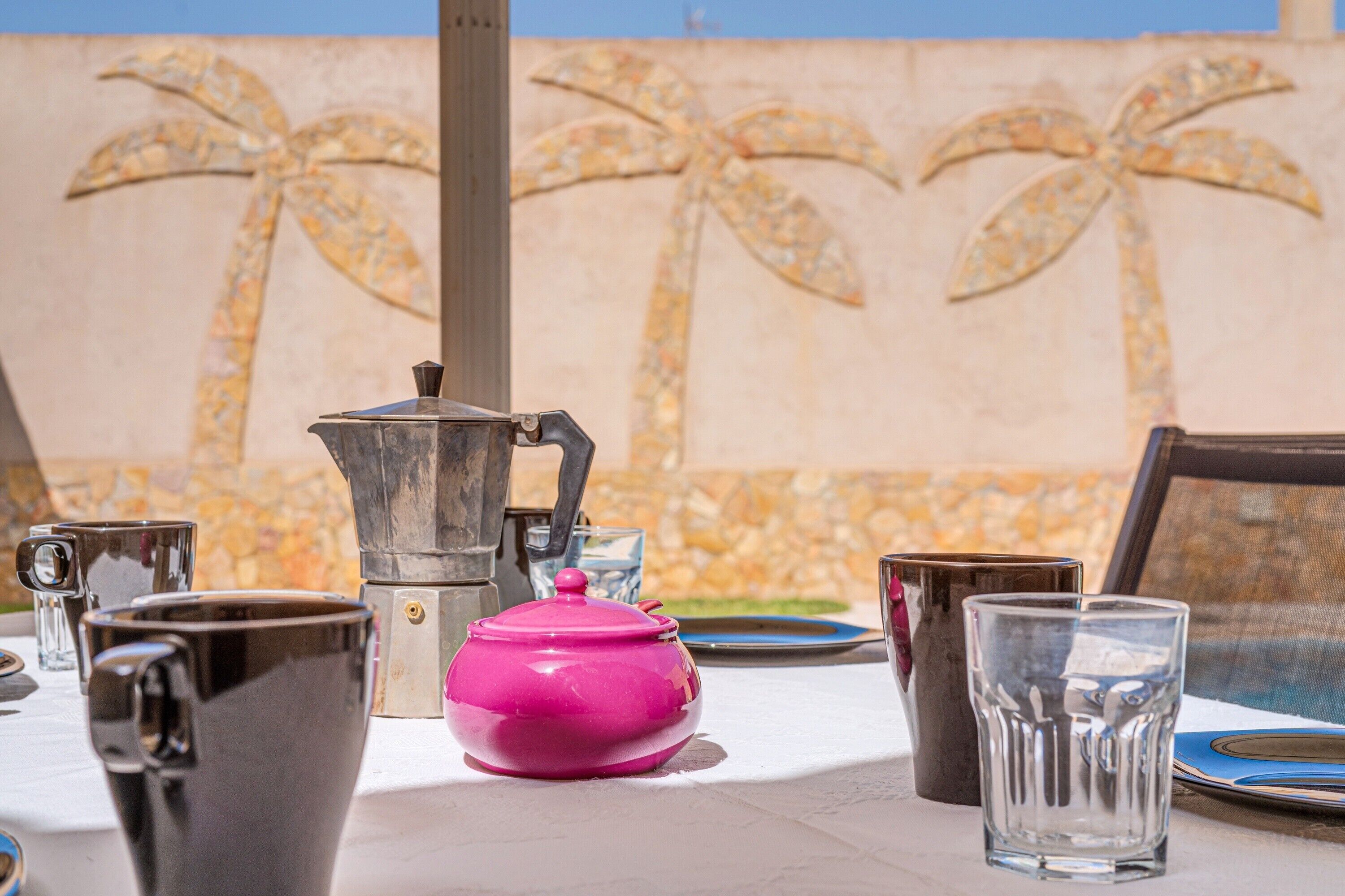 Una mesa con una cafetera, una tetera rosa y varias tazas y vasos.