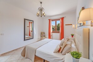 Un dormitorio con cama, candelabro, ventana con cortinas, espejo y mesita de noche con planta.