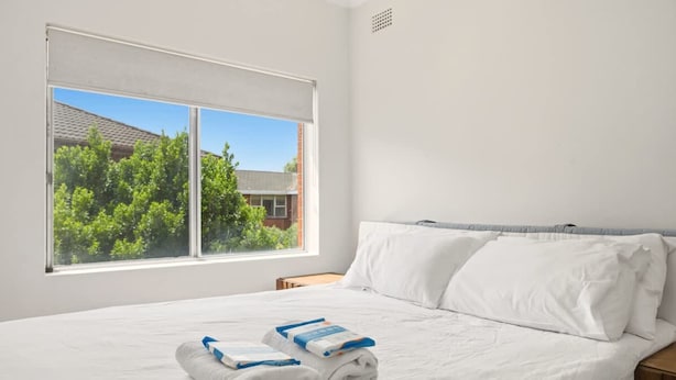 A neatly made bed with white linens and towels, a window with a view of trees and a building, and a simple wooden nightstand. - Metawise BnB 2BR Hillsdale Family Stay Sleeps 6 (Sydney)