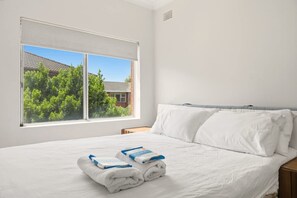 Ein ordentlich bezogenes Bett mit weißen Leinen und Handtüchern, ein Fenster mit Blick auf Bäume und ein Gebäude, sowie ein schlichter Nachttisch aus Holz.