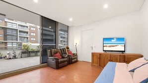 A modern living room with a large window, a leather sofa, a TV, and a wooden cabinet. - Metawise BnB Bright Open 2BR Arncliffe Sleeps 6 (Sydney)