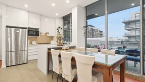 A modern kitchen with white cabinets, a stainless steel refrigerator, a microwave, and a dining area with a glass table and chairs. - Metawise BnB Bright Open 2BR Arncliffe Sleeps 6 (Sydney)