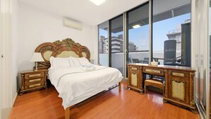 A bedroom with a large bed, wooden nightstands, a dresser, and a sliding glass door leading to a balcony with a city view. - Metawise BnB Bright Open 2BR Arncliffe Sleeps 6 (Sydney)