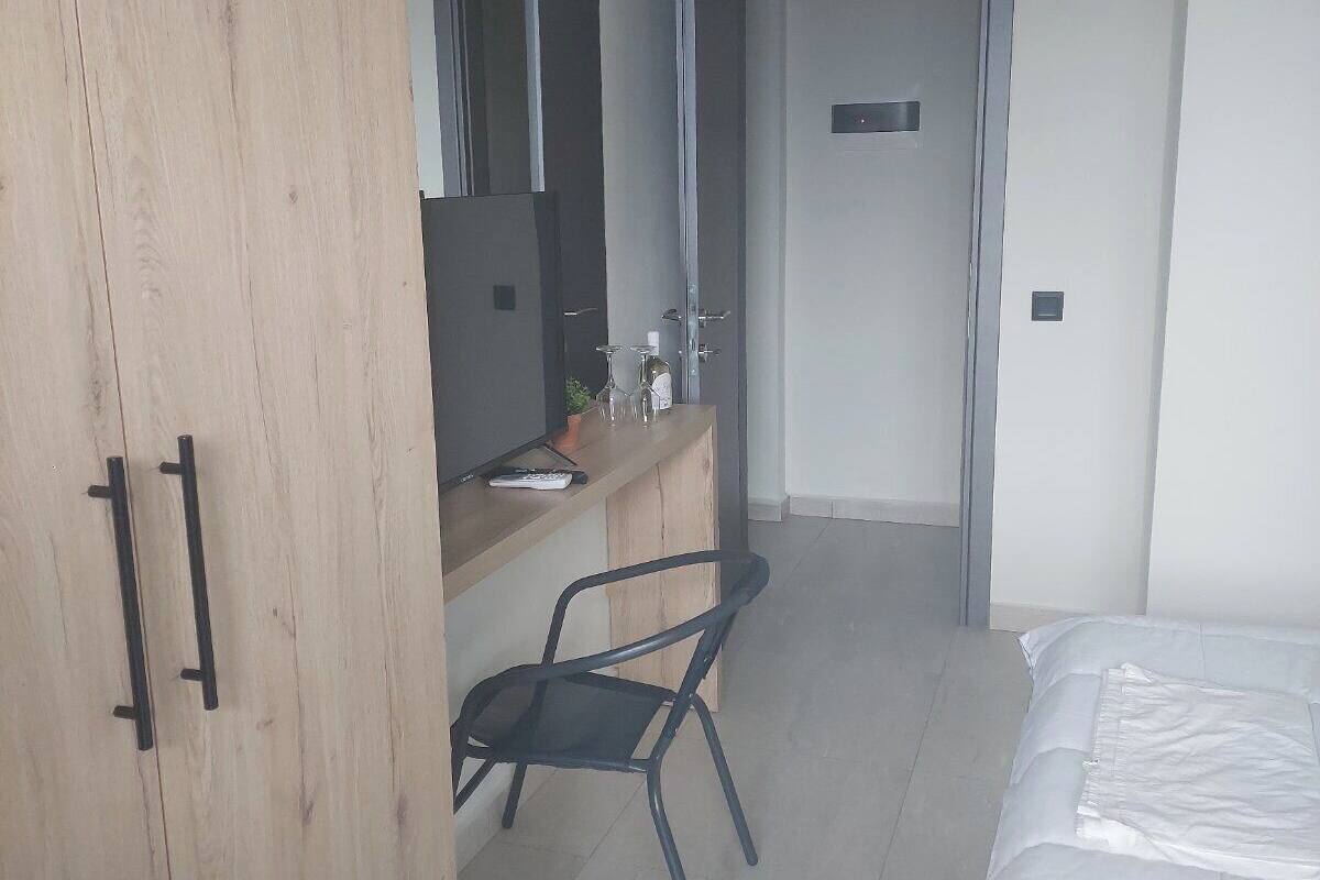 A hotel room with a wooden wardrobe, a desk with a chair, a bed, and a door leading to another room.