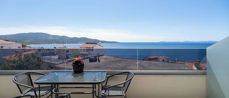 A balcony with a glass table and chairs, overlooking a coastal area with buildings and a clear sky.