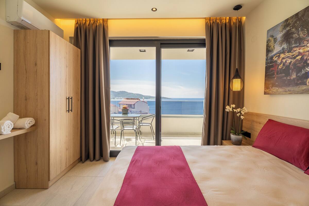 A hotel room with a bed, a wooden wardrobe, a balcony with a table and chairs, and a painting on the wall.