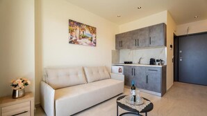 Uma sala de estar com sofá, uma mesinha com garrafa e copos, um quadro na parede e uma cozinha compacta com armários e eletrodomésticos.