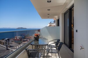 Un balcón con mesa y sillas, con vistas a la costa, edificios y un cielo azul despejado.