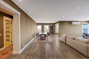 A modern living room with a wooden floor, a beige sofa, a dining table with chairs, and a view of the city through large windows. - Vesta - Luxury Nile View APT - 2BR - Maadi II (Cairo)