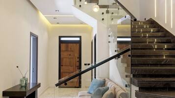 A modern interior with a marble floor, a staircase with glass railings, a sofa with cushions, and pendant lighting.