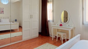 Een slaapkamer met een wit bed, een wit bureau met spiegel, een garderobe en een groot raam met gordijnen.