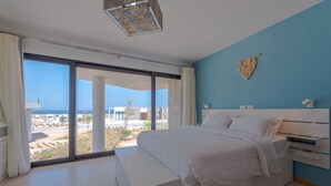 Une chambre avec un grand lit, une table de chevet et une vue sur l’océan grâce à de grandes fenêtres du sol au plafond.