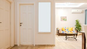 Un couloir avec une porte blanche, une grande fenêtre, un canapé à motifs floraux, une petite table et une plante en pot.
