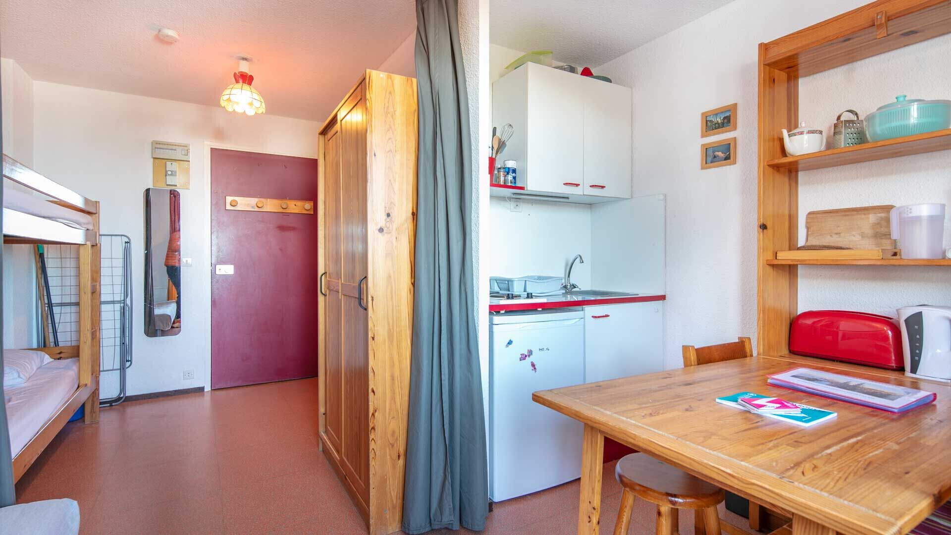 Une petite pièce comprenant un lit superposé, une porte en bois, un coin cuisine et une table à manger avec des chaises.