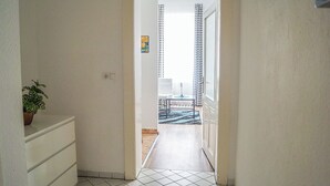 A narrow hallway with a potted plant, a small table with a lamp, and a view into a room with a patterned curtain and a chair. - Modern 2BR Apartment Long Stay Belvedere (Vienna)
