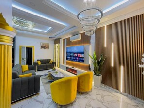 Uma sala de estar moderna com uma televisão de tela plana grande, um sofá, poltronas amarelas e uma luminária decorativa no teto.