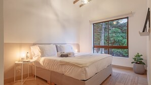 Ein modernes Schlafzimmer mit einem großen Bett, einem Nachttisch mit Lampe, einem Fenster mit Blick auf Grünflächen und einer Zimmerpflanze.