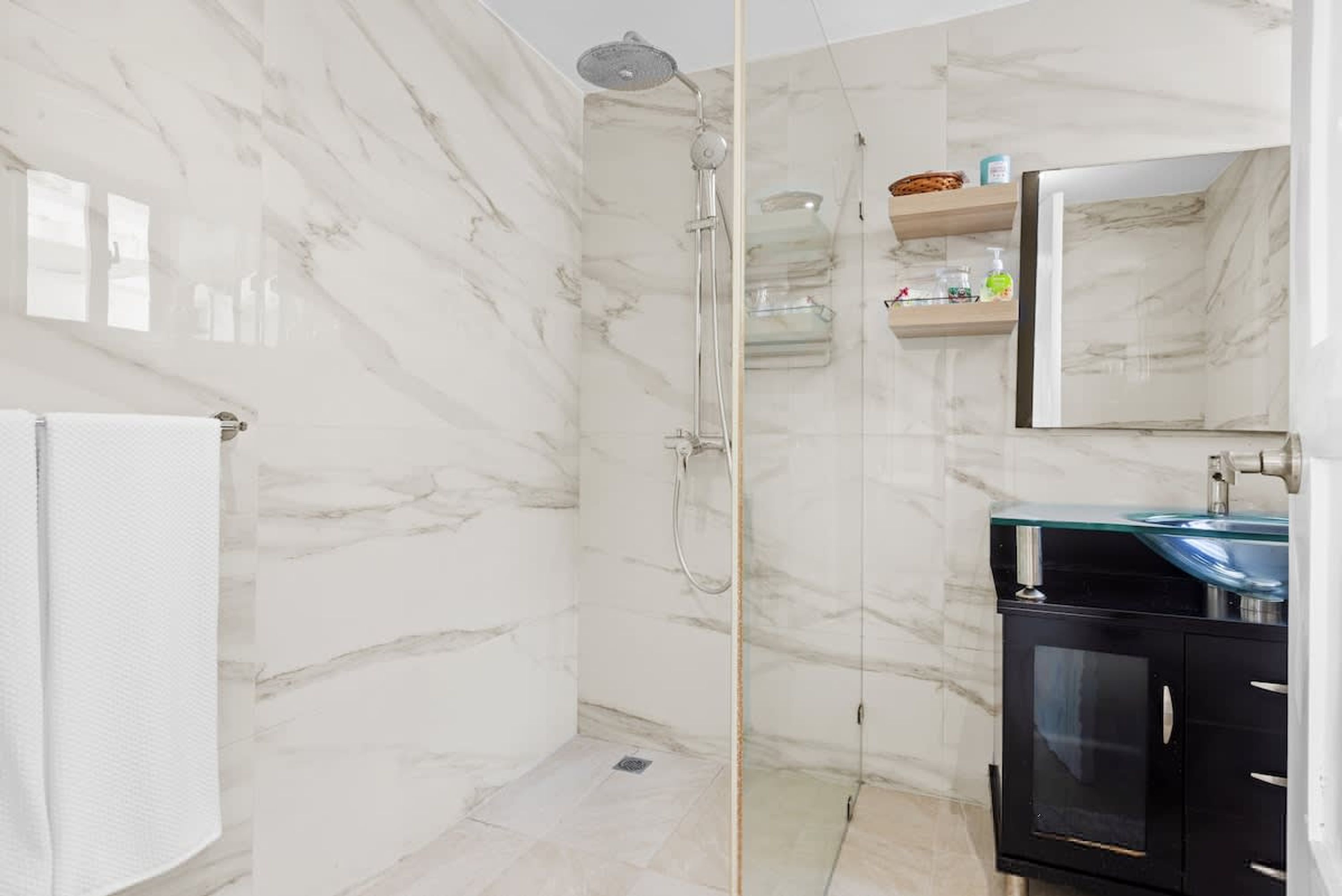 A modern bathroom with a glass-enclosed shower, marble walls, and a black cabinet with a glass countertop.