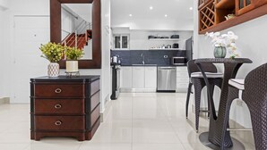 Eine moderne Küche mit weißen Schränken, einem Edelstahlkühlschrank und einer dunklen Arbeitsplatte. Es gibt einen Essbereich mit Tisch und Stühlen, sowie einen Holzschoß.