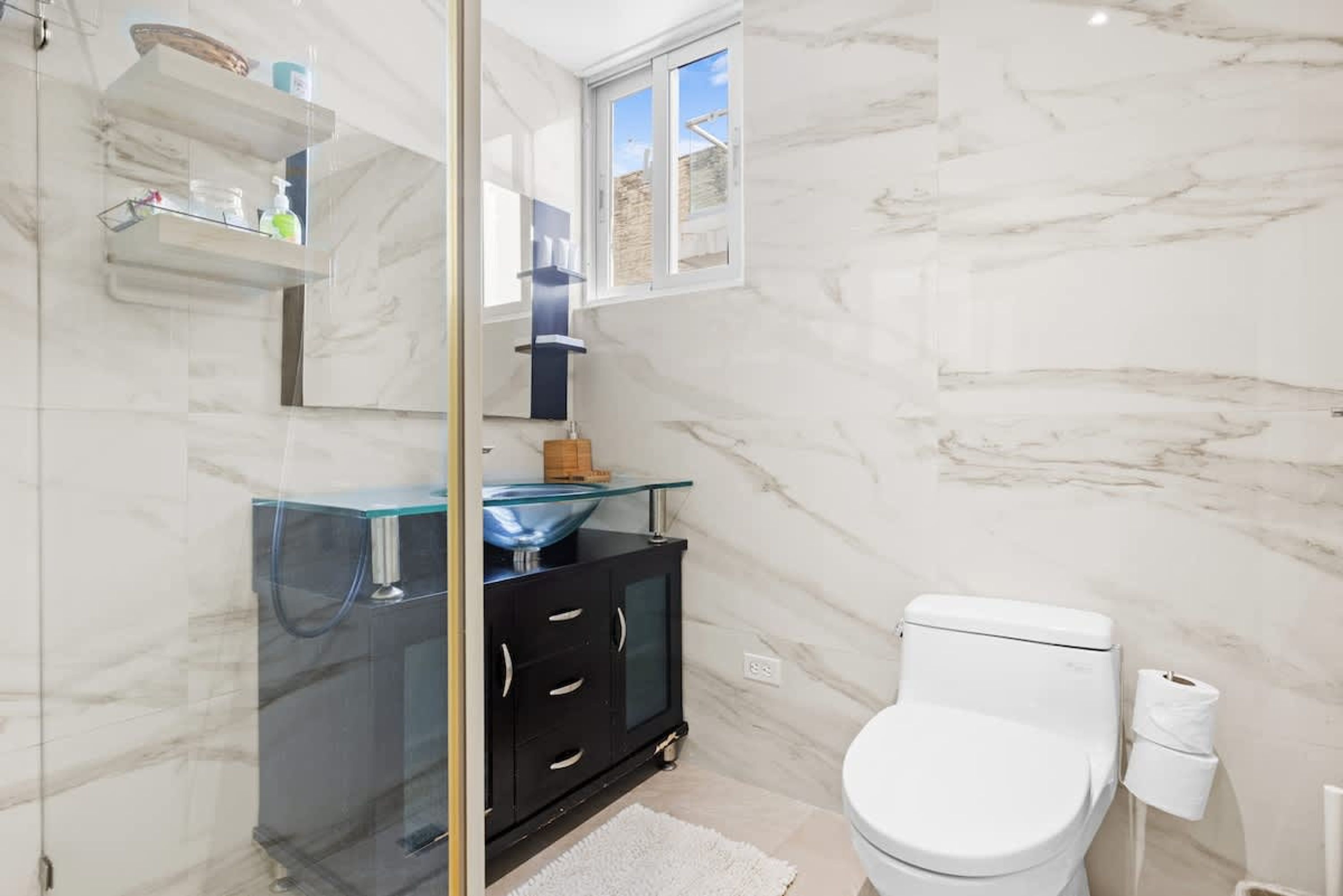 A modern bathroom with a glass shower enclosure, a black cabinet with a glass bowl sink, a white toilet, and marble walls.