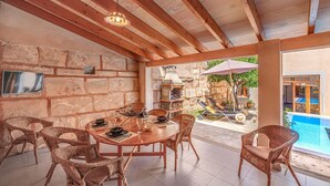 A covered patio with a wooden table and wicker chairs, a stone wall, a grill, and a pool in the background. - Beautiful Home in the Heart of Mallorca with Air Conditioning and Wi-Fi (Petra)