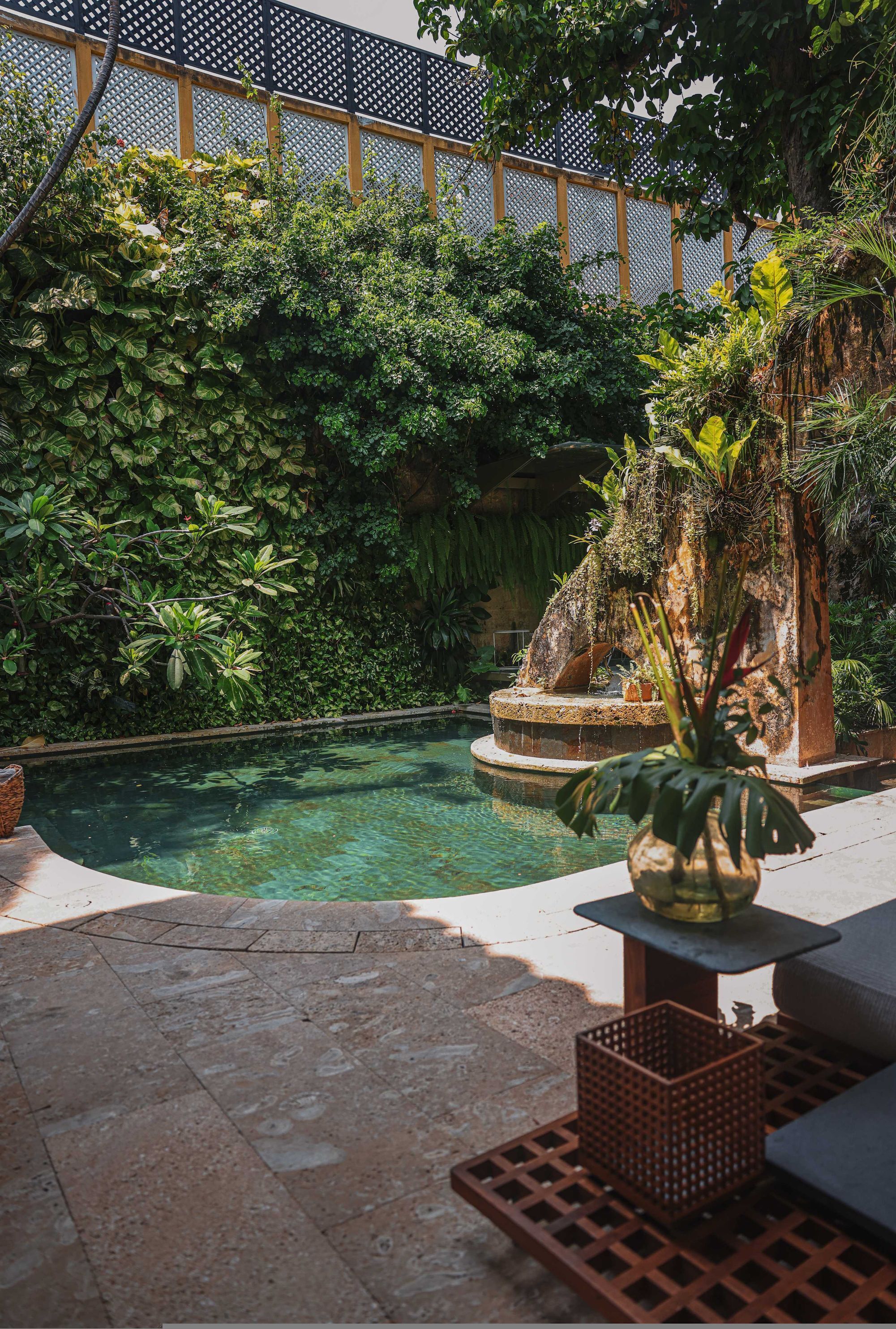 A pool surrounded by lush greenery, a stone bench, and a decorative plant.