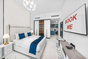 A modern hotel room with a large bed, a desk, a mirror, and a kitchen area visible through an open door. - Stayshort - Sleek and Modern Studio in Arjan That Sleeps 2 (Dubai)