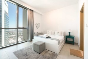 A modern bedroom with a large window, a bed with white linens, a grey ottoman, and a teal nightstand. - Stayshort - 2 Bed in Downtown Dubai That Sleeps 4 With Canal Views (Dubai)