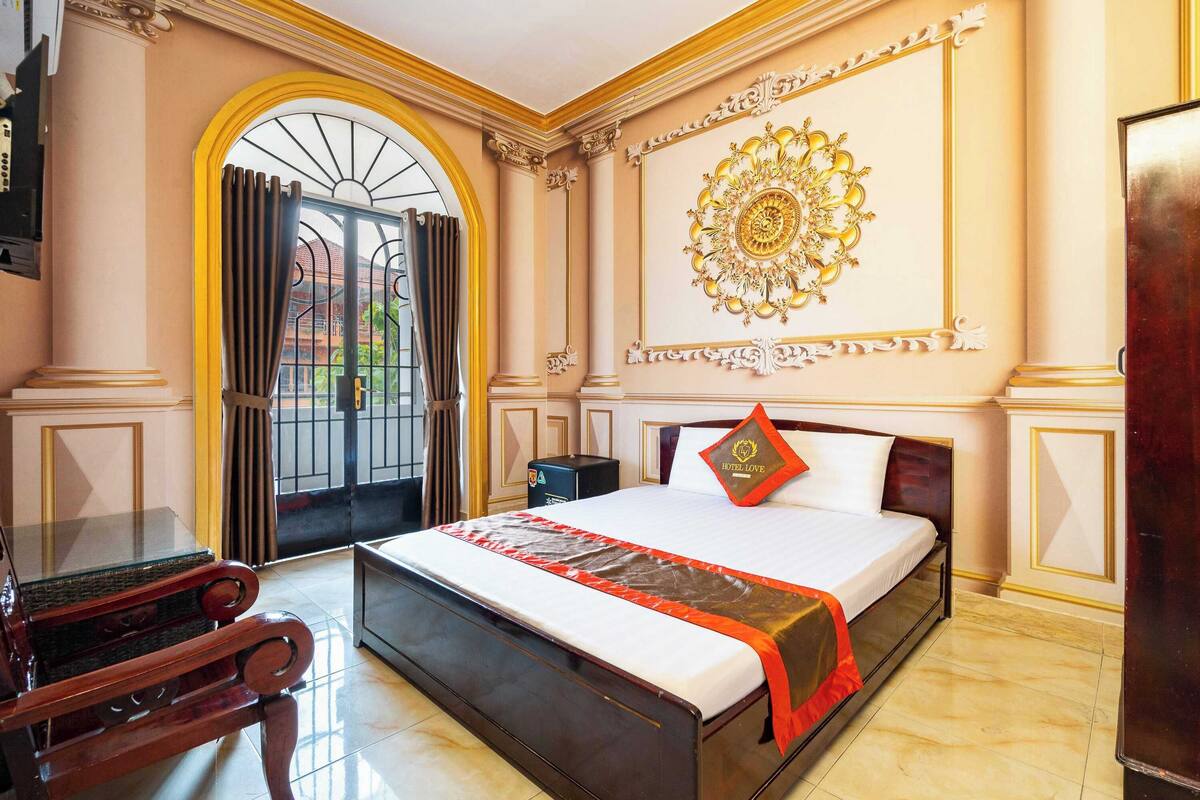 A hotel room with a large bed, a wooden chair, a decorative wall piece, and a view through a window with a metal gate.