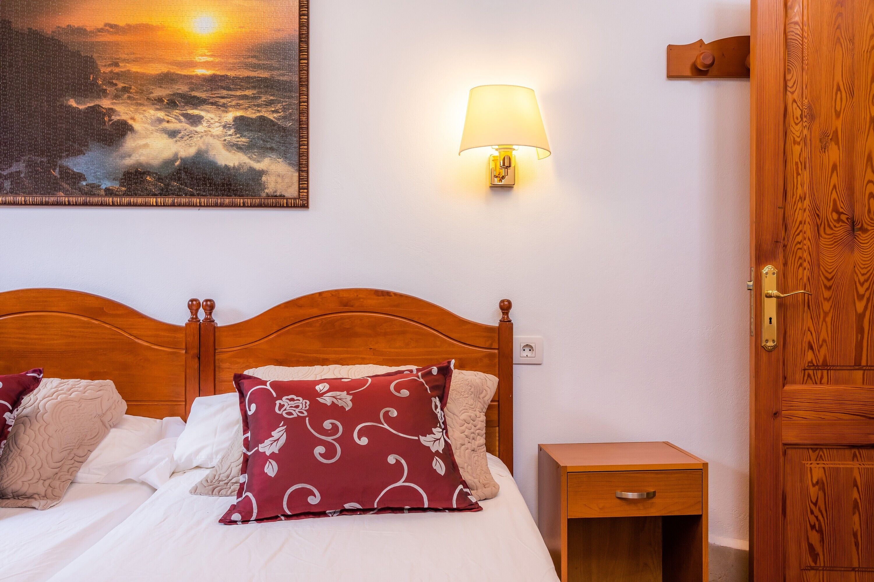 Ein Schlafzimmer mit einem Holzkopfende, einer Nachttischlampe, einem Nachttisch und einem Gemälde, das einen Sonnenuntergang über dem Meer zeigt.