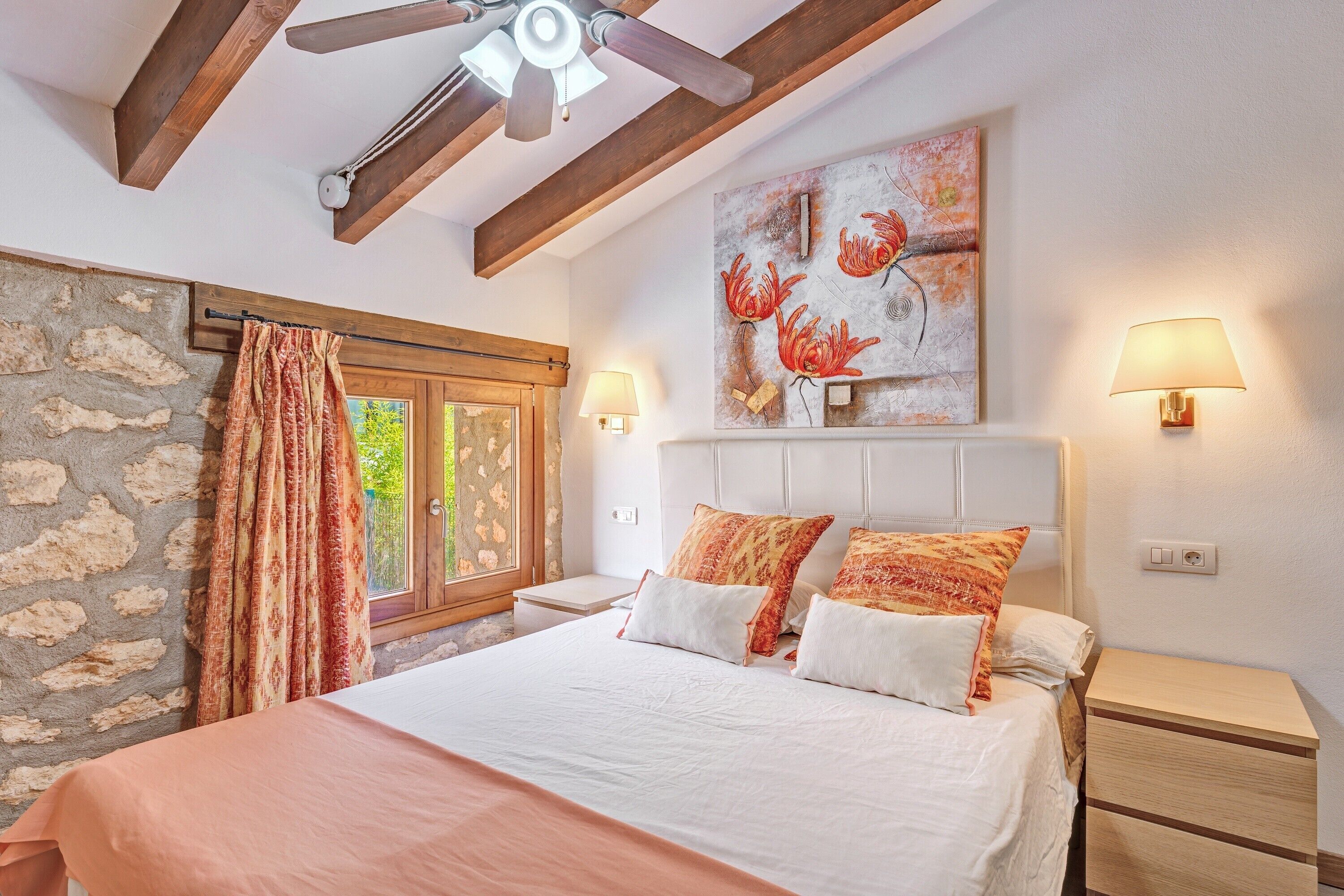 A bedroom with a stone wall, a bed with white and orange bedding, a ceiling fan, and a painting of flowers.