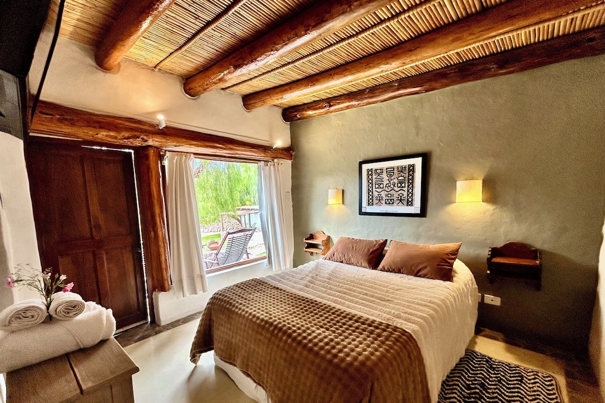 Ein Schlafzimmer mit einem großen Bett, einer Holzbalkendecke, Blick ins Freie, einer Wandleuchte und einem gerahmten Kunstwerk.