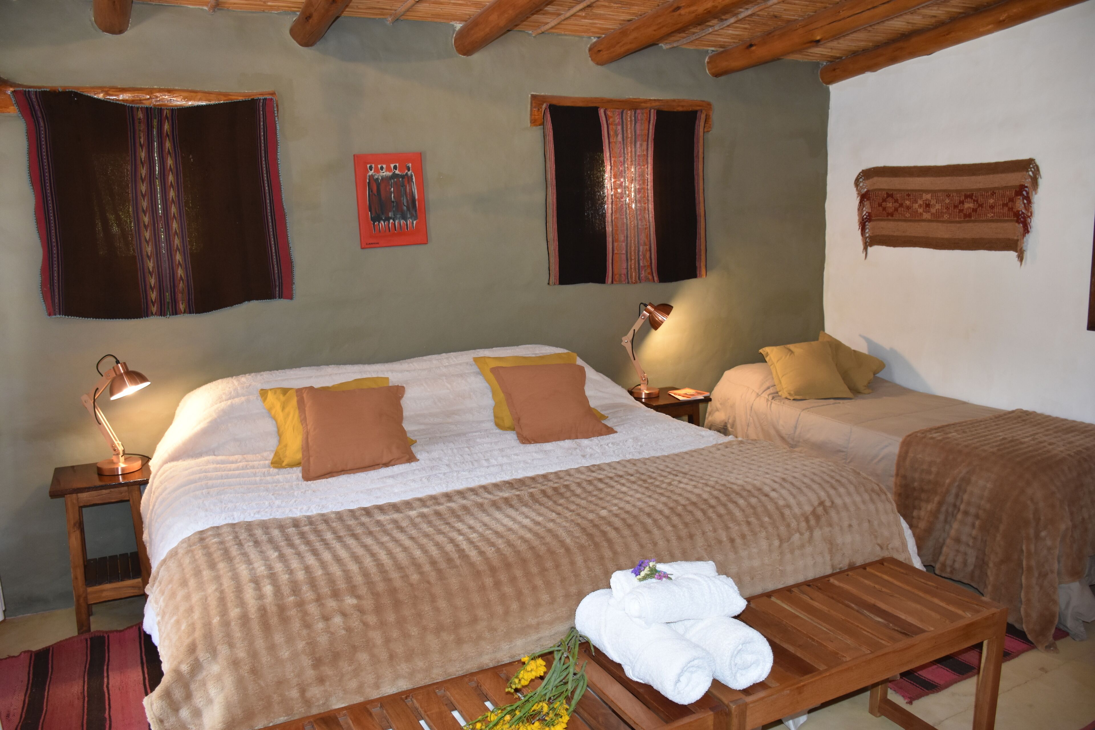 A room with a large bed, two bedside tables, a wooden bench, and a small table with towels and flowers.