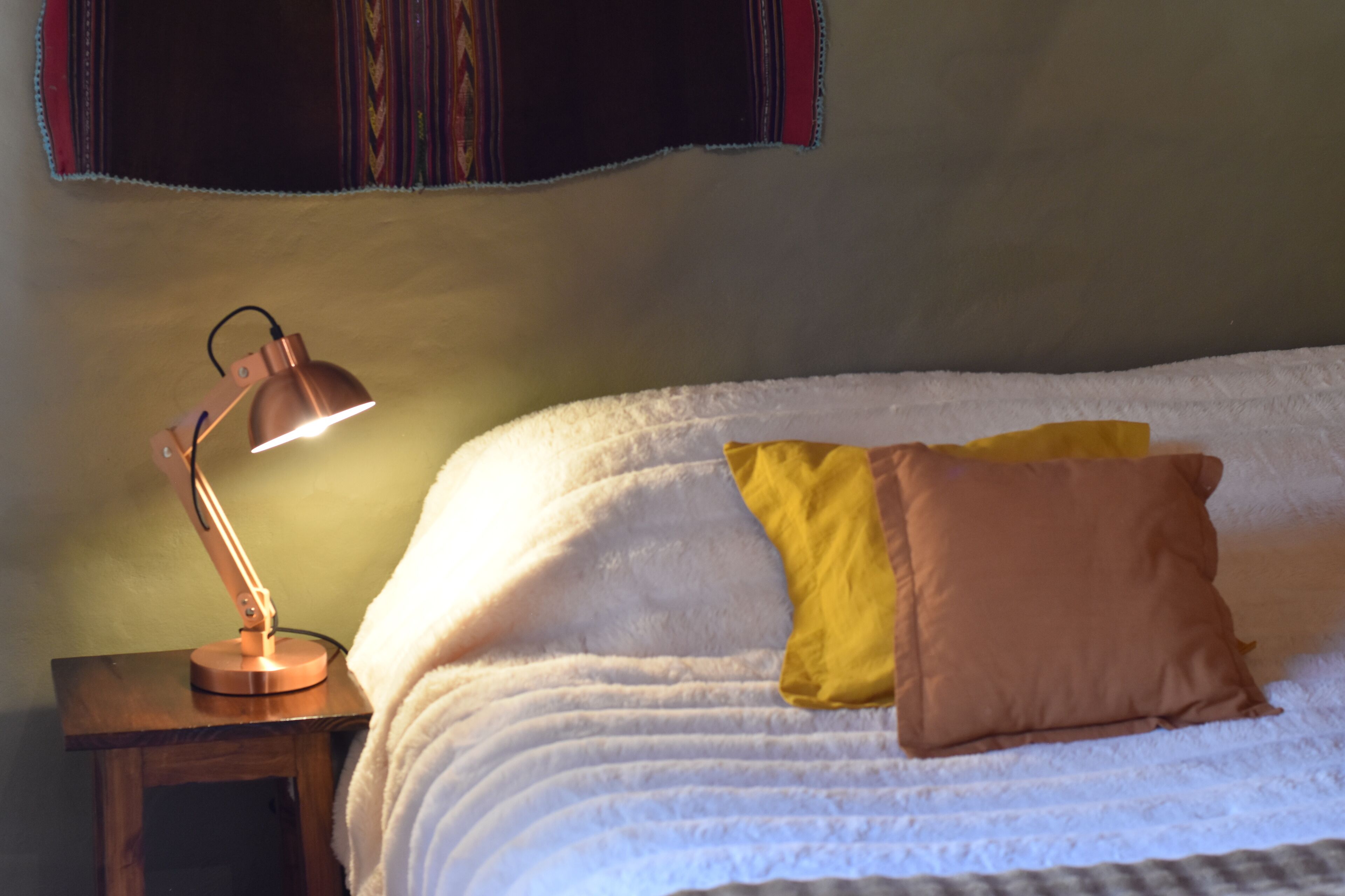 A bed with white bedding, a small wooden nightstand, a copper desk lamp, and two pillows (one yellow, one brown).