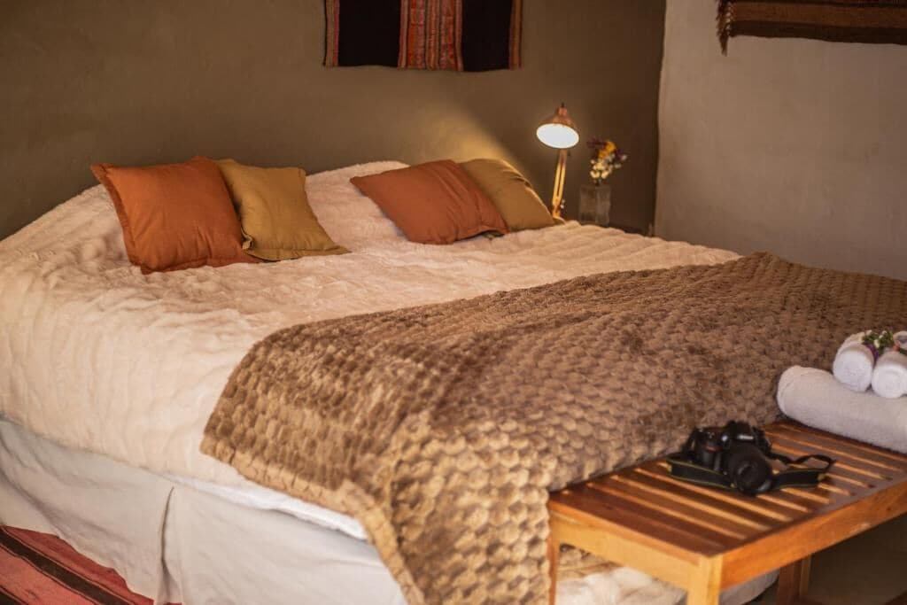 Una cama bien hecha con cubrecama blanco y almohadas marrones, una mesita de noche de madera con una cámara, y una lámpara en la mesita de noche.