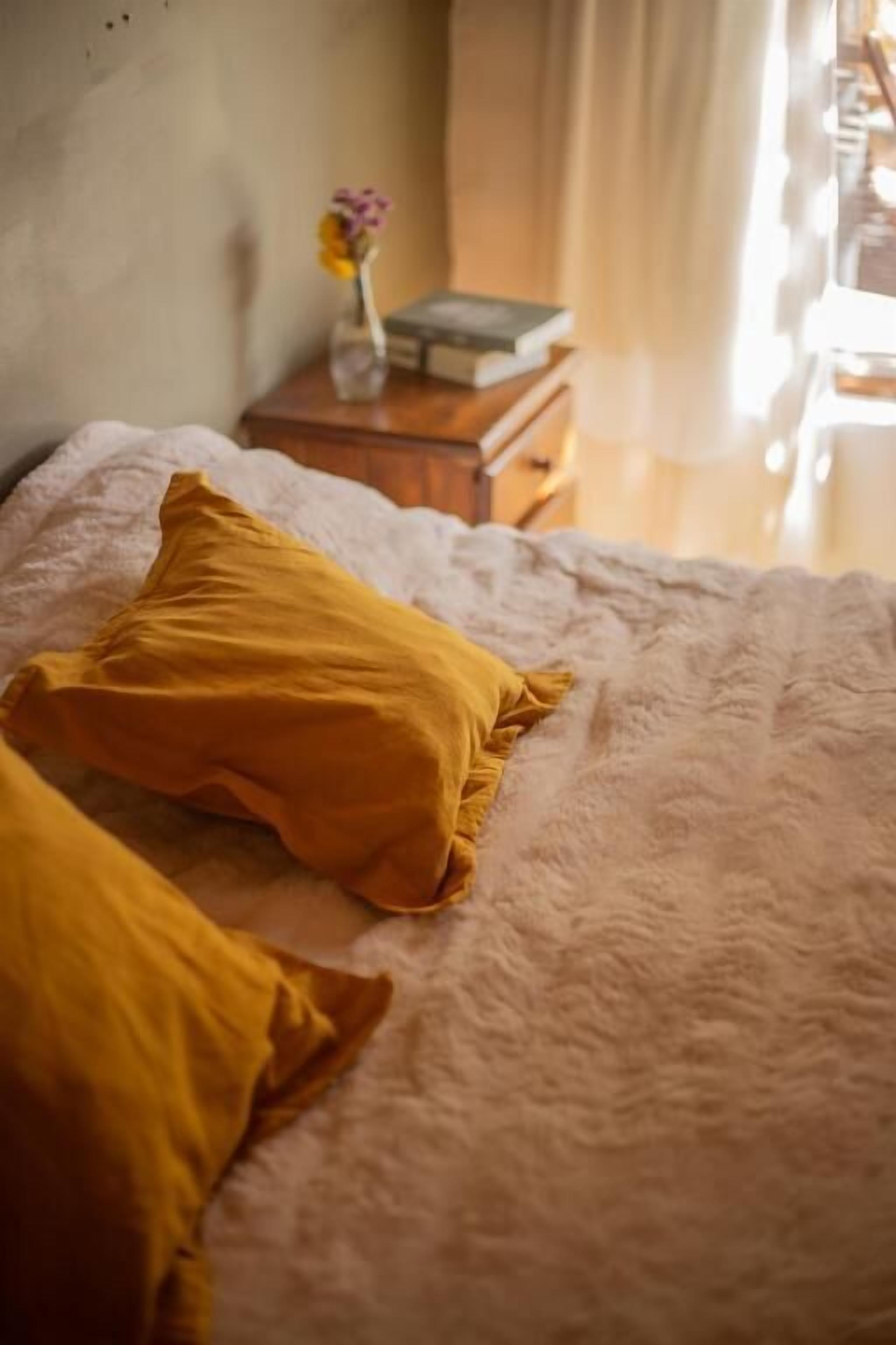 A bed with two pillows, a bedside table with a book and a vase of flowers, and a window with curtains.