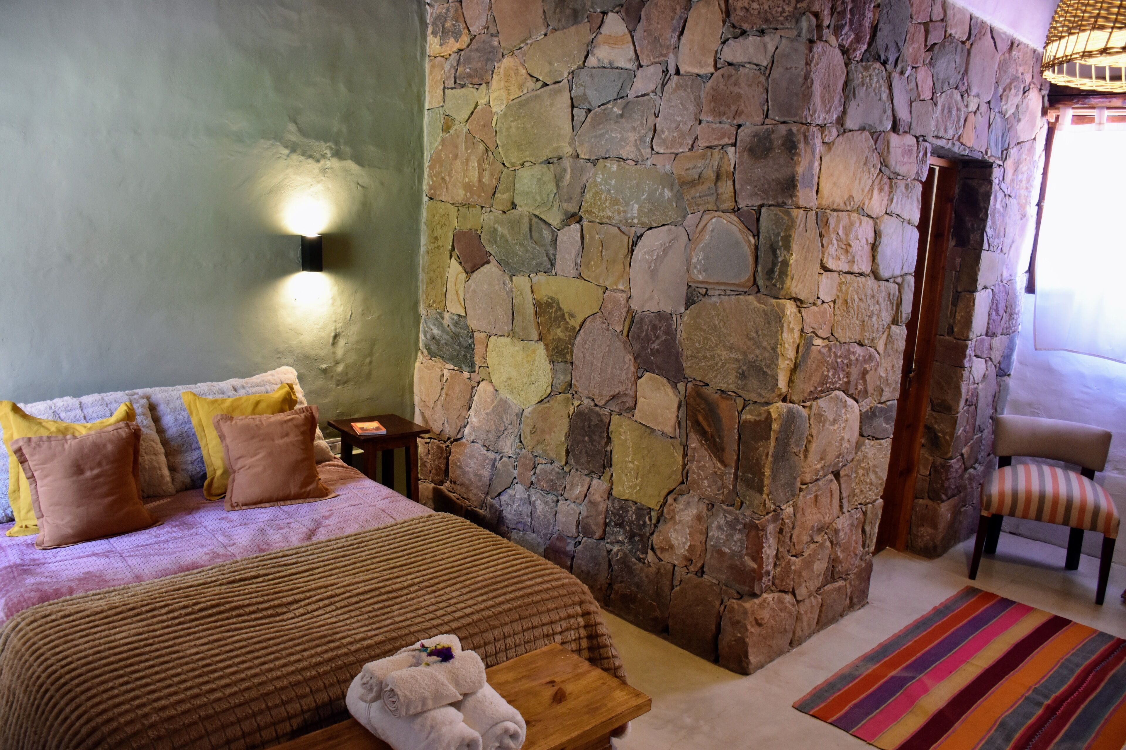 Une chambre avec un mur en pierre, un lit avec des oreillers, une table de chevet, une chaise et un tapis à rayures.