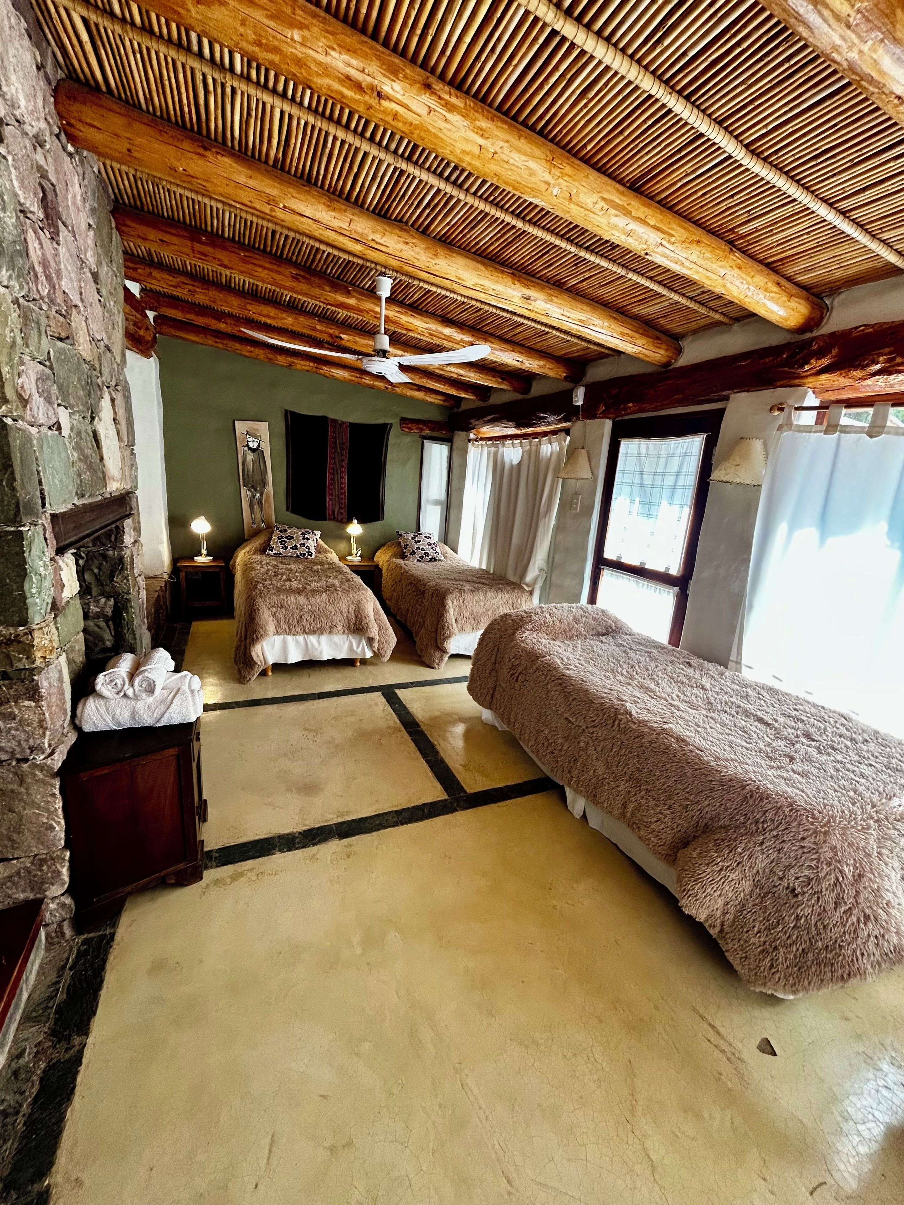 A room with two beds, a stone wall, a wooden ceiling, and a large window.