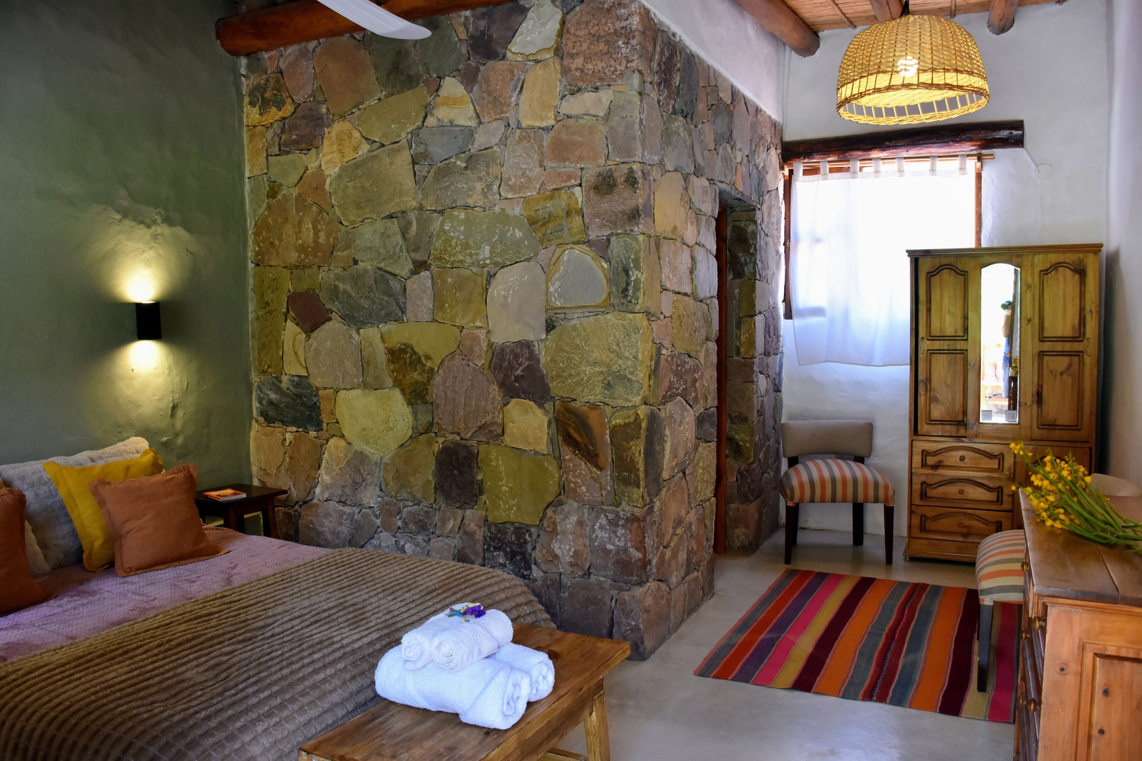 A bedroom with a stone wall, a wooden bed, a bench, a wooden dresser, and a mirror.